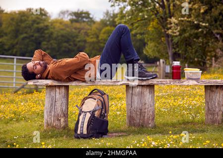 Mann, Der Auf Einer Bank Auf Dem Land Liegt, Entspannt Sich Und Musik Oder Podcast Über Kabellose Kopfhörer Hören Stockfoto