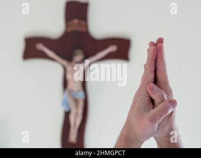 Nahaufnahme von zwei verbundenen Händen im Gebet. In einem hellen Hintergrund ein verschwommenes Kruzifix. Stockfoto