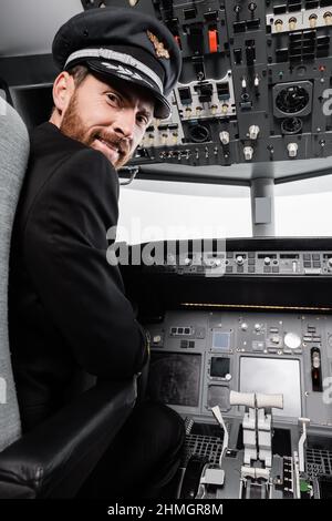 Bärtiger Pilot mit Mütze lächelt und schaut in einem Flugsimulator auf die Kamera Stockfoto
