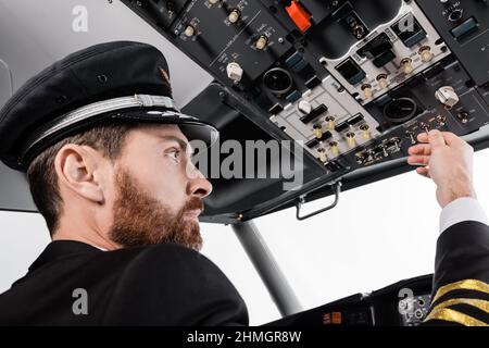 Bärtiger Pilot in der Kappe, der die Deckenplatte mit einem Satz von Schaltern im Flugsimulator erreicht Stockfoto