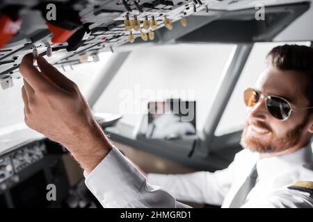 Verschwommener bärtiger Pilot mit Sonnenbrille, der über die Decke reicht und im Flugsimulator lächelt Stockfoto