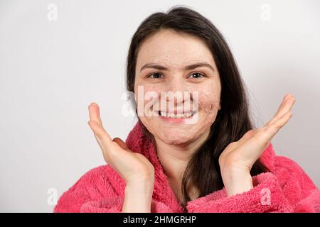 Eine junge Brünette Frau in einem rosa Bademantel macht reinigende Gesichtsbehandlungen - eine Maske oder Peeling, um die Haut zu reinigen und zu nähren. Stockfoto