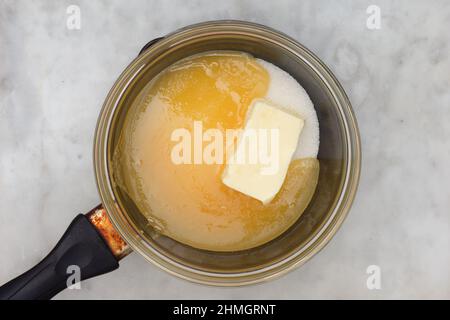 Draufsicht auf Bain-marie mit Zucker, Butter und Honig in Glasschüssel auf Kochtopf als Zutaten von Kuchen medovik auf Marmorboden Stockfoto