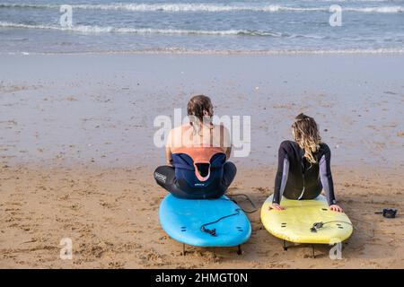 Zwei Surfer machen eine Pause und sitzen auf ihren Surfbrettern an der Küste am Fistral Beach in Newquay in Cornwall in Großbritannien. Stockfoto