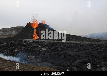 Eruptionsausbruch beim Vulkanausbruch Fagradalsfjall, Island. Verkrustete schwarze Lava steht im Vordergrund und Dampf steigt auf. Stockfoto