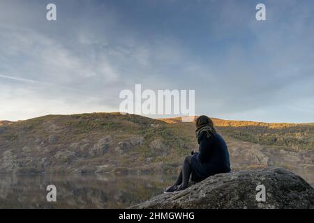 Frau, die auf einem großen Felsen am Rand eines Gletschersees sitzt. Sanabria Lake. Stockfoto