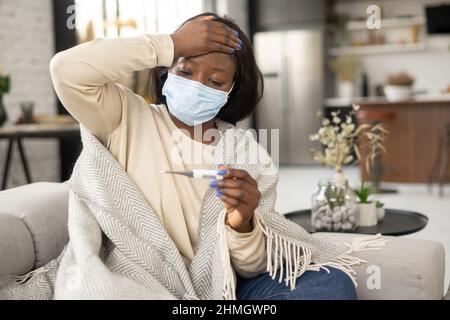 Krank verärgert afroamerikanische Frau trägt Maske Messung Temperatur sitzen zu Hause. Kranke schwarze Dame mit Erkältung oder Grippe schält Fieber und leidet an Viruserkrankungen, Selbstisolationskonzept Stockfoto