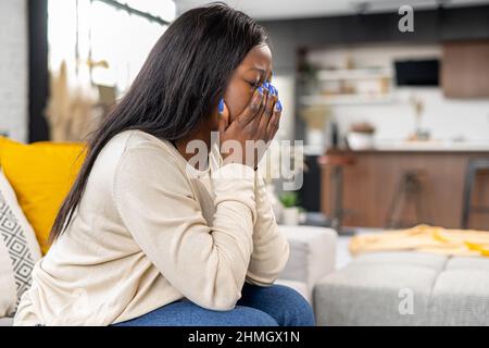 Verstörte afroamerikanische Frau, die in Trauer auf dem Sofa sitzt, die Augen mit Handflächen schließt und weint, enttäuschte Frau fühlt Verzweiflung, Besorgnis, Depression, Einsamkeit, leidet unter starken Kopfschmerzen Stockfoto