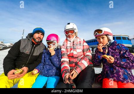 Die Familie genießt das Mittagessen während einer Pause im Urlaub in Skiausrüstung. Familie mit Kindern auf dem Skiurlaub essen Sandwiches in Helmen und Schutzbrillen. Winter Stockfoto