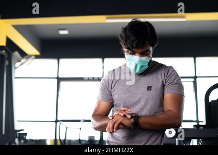 Junge Muskelaufbau mit medizinischer Gesichtsmaske Überprüfung Trainingsplan oder Überwachung hart-Rate von Smartwatch im Fitnessstudio - concpet der Gesundheitsversorgung, Technologie Stockfoto