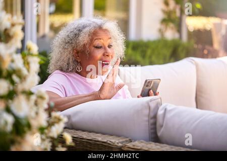 Ältere Frau Im Ruhestand, Die Draußen Im Garten Zu Hause Sitzt Und Videoanrufe Auf Dem Mobiltelefon Führt Stockfoto