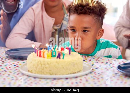 Familie, Die Zu Hause Am Tisch Sitzt Und Feiert, Wie Ein Junge Kerzen Auf Dem Geburtstagskuchen Bei Der Party Ausbläst Stockfoto