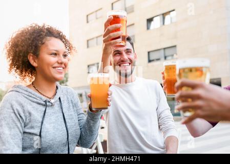 Eine Gruppe von glücklichen Freunden klirren auf Biergläsern, während sie auf der Terrasse des Cafés in der Stadt sitzen und Zeit miteinander verbringen Stockfoto