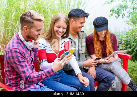 Eine Gruppe von multikulturellen, glücklichen Freunden, die ihre Handys auf einer leuchtend roten Parkbank im Freien benutzen, teilen Medien, lachen und scherzen gemeinsam in einem Hig Stockfoto