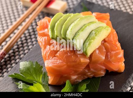 Von oben frischer roher Lachskartar mit Scheiben Avocado serviert auf schwarzem Brett mit Petersilie in der Nähe von Essstäbchen in hellem Café Stockfoto