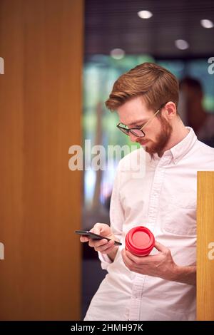 Geschäftsmann In Der Pause Im Büro Hält Wiederverwendbare Kaffeetasse Und Blick Auf Handy Stockfoto