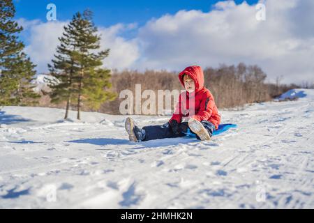 Fröhlicher und positiver kleiner Junge, der Rodeln und kaltes Wetter im Freien genießt, Spaß im Winter, Aktivitätskonzept Stockfoto