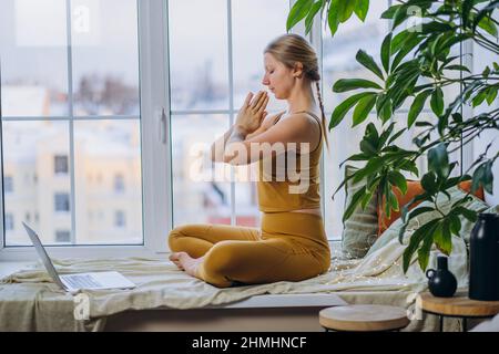 Die junge blonde langhaarige Frau in gelber Farbe meditiert in Lotuspose und sitzt auf einer Decke, die das Fensterbrett bedeckt, und schaut auf den Laptop-Bildschirm Stockfoto