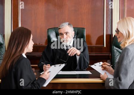 Senior Richter hält Brillen in der Nähe von Anwalt und Staatsanwalt Schreiben auf Klemmbrettern Stockfoto
