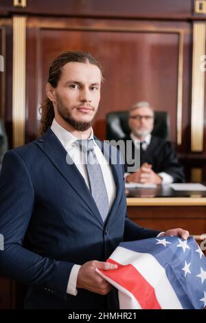 Anwalt, der die Kamera anschaut, während er die us-Flagge in der Nähe des verschwommenen Richters im Gerichtssaal hält Stockfoto