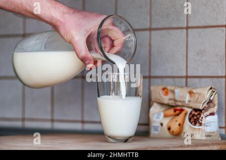 Die Hand eines Mannes hält einen Krug und gießt Milch in ein Glas. Eine Packung Kekse verschwimmt im Hintergrund Stockfoto