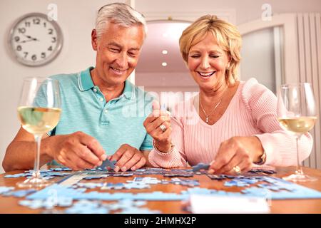 Ältere Pensionierte Paar Sitzt Am Tisch Zu Hause Mit Einem Glas Wein Tun Puzzle Stockfoto
