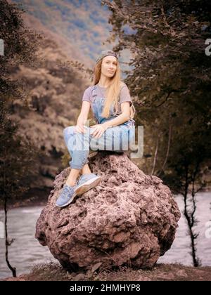 Lächelnde blonde Frau in Denim-Overalls sitzt auf einem runden großen Stein im Waldtal vor dem Hintergrund eines Flusses Stockfoto