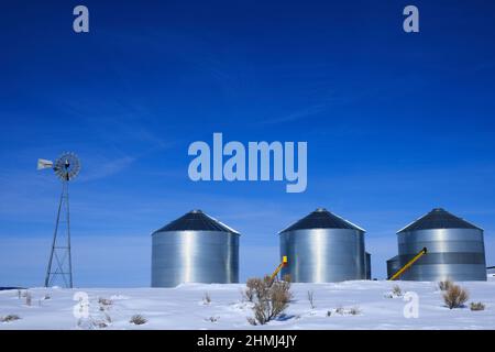 Windmühle und Stahlkornsilos im Winterschnee mit blauem Himmel auf dem Bauernhof für die landwirtschaftliche Landwirtschaft Stockfoto