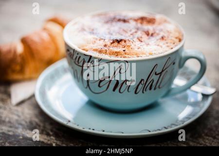 Heiße Schokolade mit einem Prise Kakaopulver in einem dekorativen Becher mit einem Croissant auf einer rustikalen Holzarbeitsfläche Stockfoto