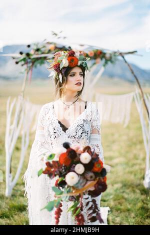 Die Braut in einem Kranz mit einem Blumenstrauß steht neben dem Hochzeitbogen Stockfoto