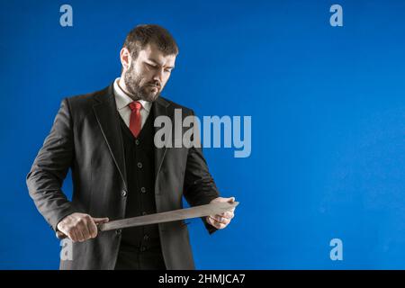 Ein bärtiger Mann in einem Anzug mit einer roten Krawatte und einem langen Messer in den Händen. Der Mörder untersucht sein Messer. Gefährlicher Mann. Bedrohung des Lebens. Stockfoto