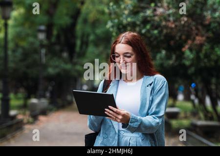 Junge Geschäftsfrau mit Rotschopf in blauem Anzug, die mit Geschäftspartnern im Freien auf einem digitalen Tablet spricht, professionelle Architektin Stockfoto