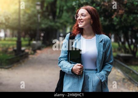 Attraktive junge Geschäftsfrau mit blauem Anzug, der ein digitales Tablet im Freien hält, professionelle Architektin, die im Park spazieren geht, mit Lichtreflexen Stockfoto