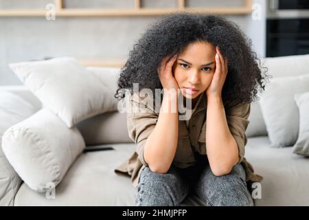 Frustrierte afroamerikanische Frau sitzt in Trauer auf dem Sofa, enttäuschte multirassische Frau fühlt Verzweiflung, Besorgnis, Depression, Einsamkeit, leidet unter starken Kopfschmerzen Stockfoto