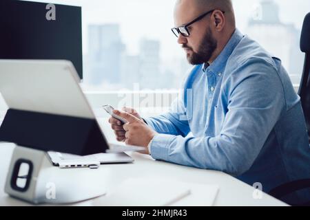 Ernsthafter männlicher Manager, der während konzentrierter Arbeit im Büro auf dem Smartphone surft Stockfoto