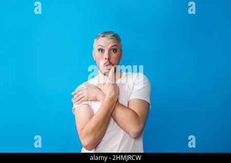 Ein schwuler Mann mit Make-up in einem weißen T-Shirt auf blauem Hintergrund, der auf die Kamera schaut. Das Konzept von LGBT Stockfoto