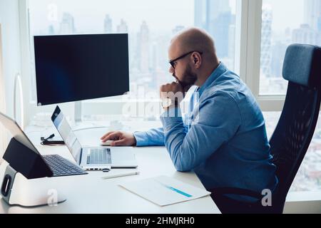 Nachdenklicher junger intelligenter Geschäftsmann, der während der Arbeit im Büro Netbook durchstöbert Stockfoto