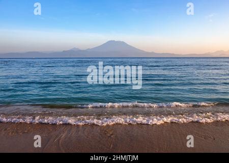 Aktiver Vulkan Mount Agung in Nusa Penida, Bali, Indonesien. Toller Sonnenuntergang. Sandstrand. Hohe Wellen mit Schaum breiten sich an der Küste aus. Stockfoto