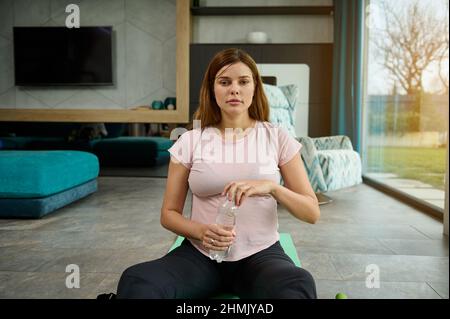 In Erwartung eines Babys sitzt die junge Frau auf einer Yogamatte in Lotuspose und trinkt Wasser, während sie sich nach einem leichten Training zu Hause entspannt. Schwangerschaft und Sport Stockfoto