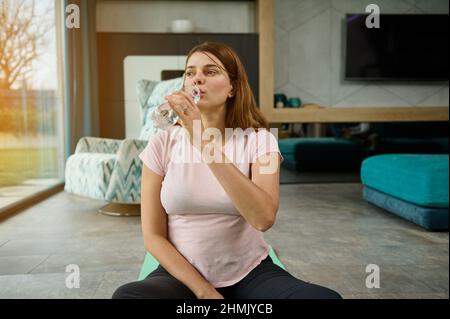 Die hübsche Frau sitzt in Erwartung eines Babys auf einer Yogamatte in Lotuspose und trinkt nach einem leichten Training zu Hause Wasser. Schwangerschaft, Fitness und Sport Konz Stockfoto