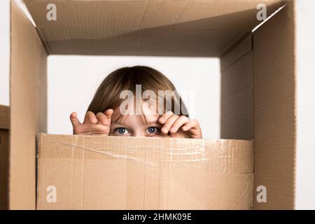 Aufgeregter kleiner Junge, der in einen riesigen Karton springt. Er spielt und schaut aus einer Box. Kid freut sich über den Umzug in ein neues Zuhause. Stockfoto