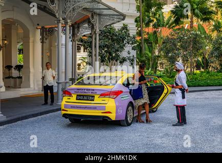 Singapur - 08. September 2019: Schwarze Frau, die am berühmten Hotel des Rafales aus dem Taxi tritt, unterstützt vom Portier Sikh in einer Militäruniform Stockfoto