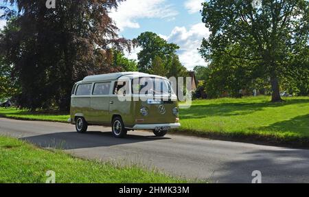 Klassischer Volkswagen Camper Van, der durch Dorfhaus und Bäume im Hintergrund fährt. Stockfoto