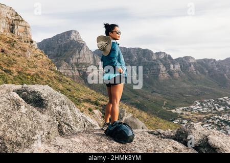 Junge Frau in Sportkleidung mit Rucksack, die auf die Aussicht schaut, während sie auf einem Felsen steht Stockfoto