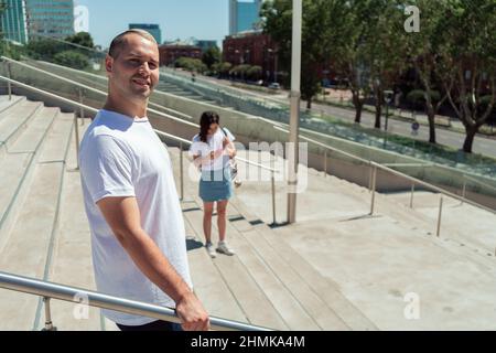 Ein Mann, der sich an einem Geländer festhält, während er eine Treppe hinuntergeht, und eine unfokussiere Frau im Hintergrund mit ihrem Handy. Stockfoto