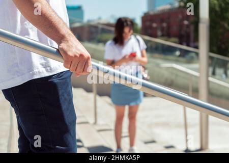 Ein nicht erkennbarer Mann, der sich an einem Geländer festhält, während er eine Treppe hinuntergeht, und eine unfokussiere Frau im Hintergrund mit ihrem Mobiltelefon. Stockfoto