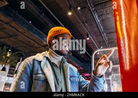 Ein positiver afroamerikanischer Mann mit orangefarbenem Strickhut und kabellosen Kopfhörern bestellt Essen über einen Selbstbedienungskioské im Café Stockfoto