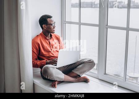 Der attraktive afroamerikanische Mann in legerer Kleidung und Brille surft auf einem modernen Laptop, der zu Hause auf der Fensterbank sitzt, ins Internet Stockfoto