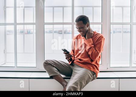 Positiver afroamerikanischer Mann in stilvollem orangefarbenem Hemd sieht aus dem Fenster und lächelt mit einem modernen Mobiltelefon in einem hellen Raum zu Hause Stockfoto