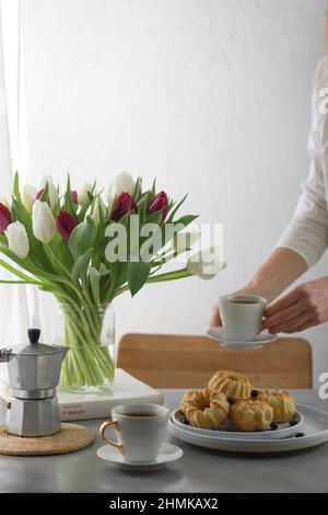 Mini-Bunt-Kuchen mit Früchten auf dem Teller, zwei Tassen Kaffee und Banch Tulpenblumen in einer Vase. Mädchen hält eine Tasse Kaffee. Stockfoto